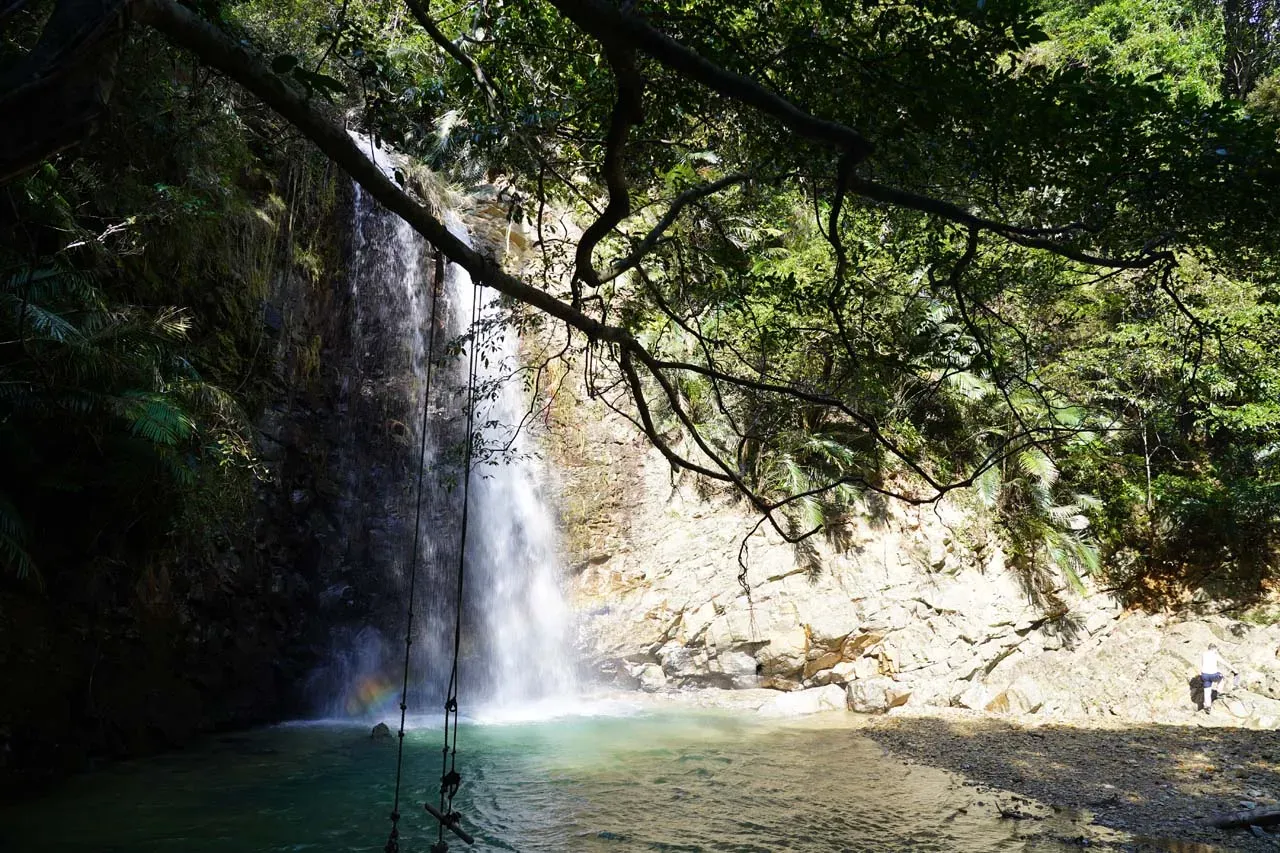 Person standing on rocks near Taataki Waterfall base with rainbow in mist at Taataki Waterfall Japan
