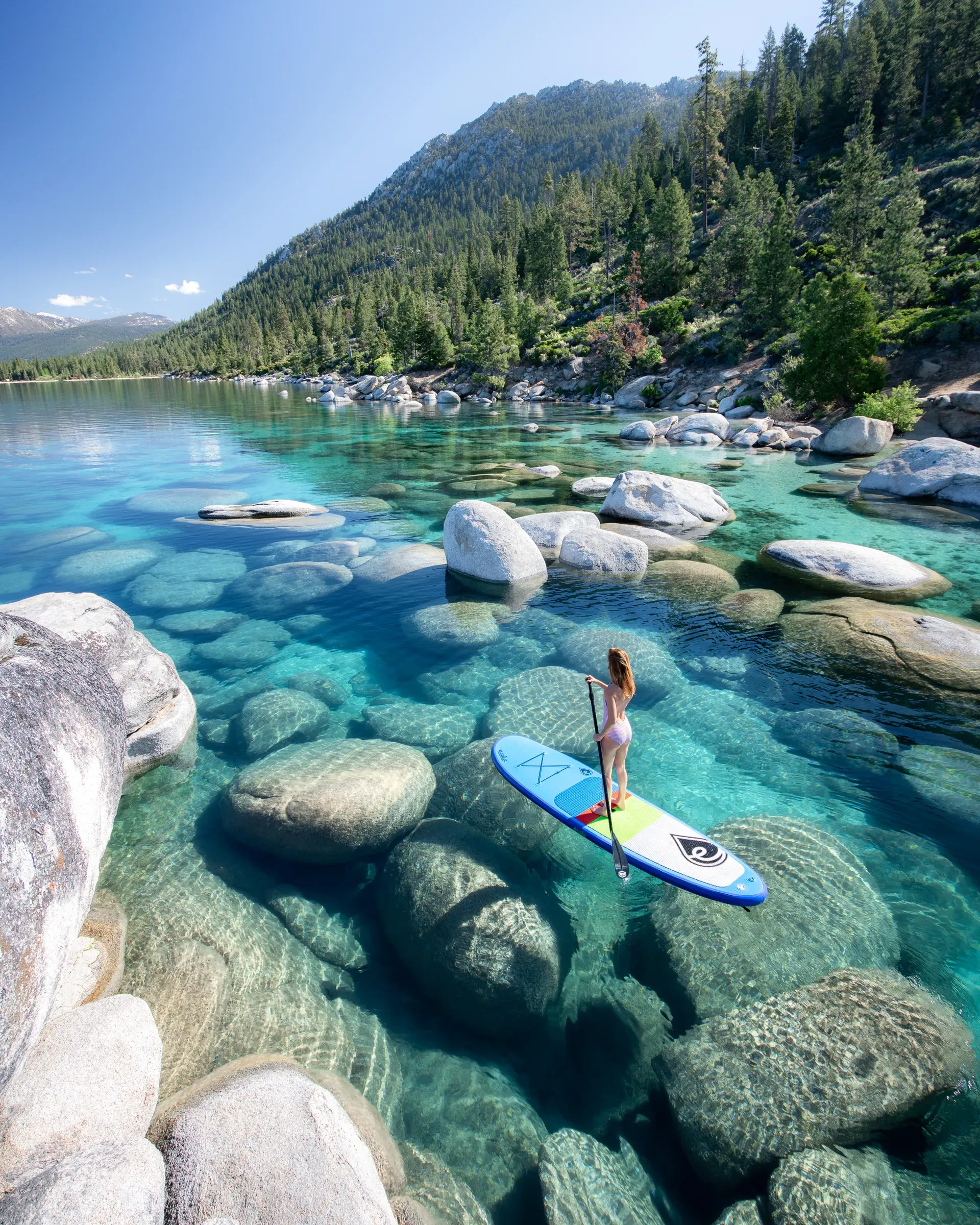 Woman stand-up paddleboarding on the crystal clear turquoise waters of Lake Tahoe surrounded by smooth rocks