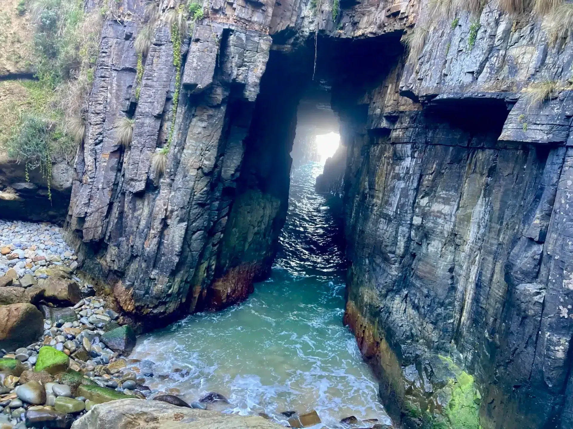 A narrow rocky water channel runs through Remarkable Cave illuminated by sunlight streaming from the opening