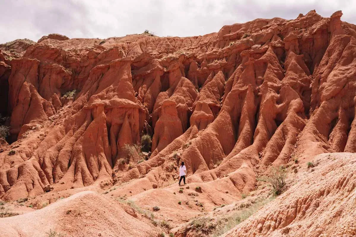 Person standing next to towering reddish sandstone formations with sharp rugged peaks in Skazka Canyon Kyrgyzstan