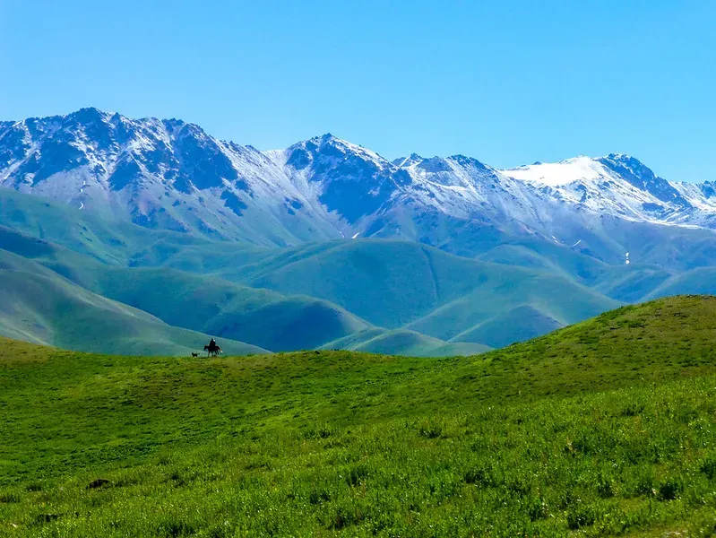 Kyrgyz horseman riding across summer pastures near Song Kol Lake with snow-capped Tian Shan peaks behind