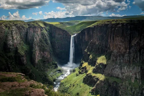 A tall narrow waterfall plunges into a deep lush green canyon with rugged cliffs under a partly cloudy sky in Lesotho