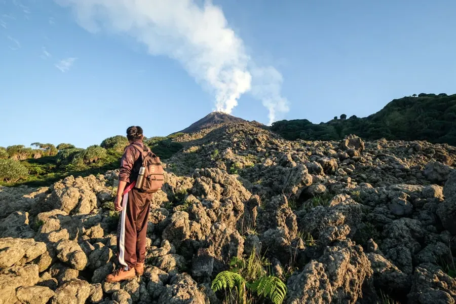 Hiker with backpack on rocky terrain looking up at Mount Karangetang's smoking volcanic cone on Siau Island, Indonesia