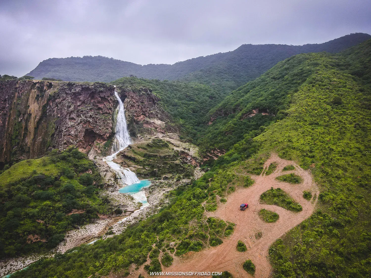 Aerial perspective of Wadi Darbat waterfall and turquoise lake surrounded by green hills and rocky cliffs in Dhofar, Oman
