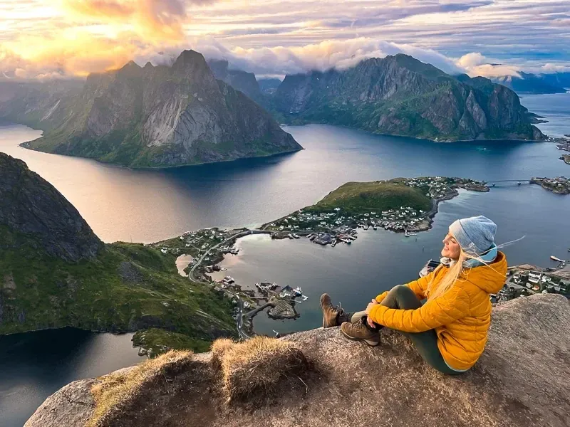 Hiker sitting on a rocky summit ledge at Reinebringen gazing at sunset over Reinefjord and the Lofoten Islands, Norway