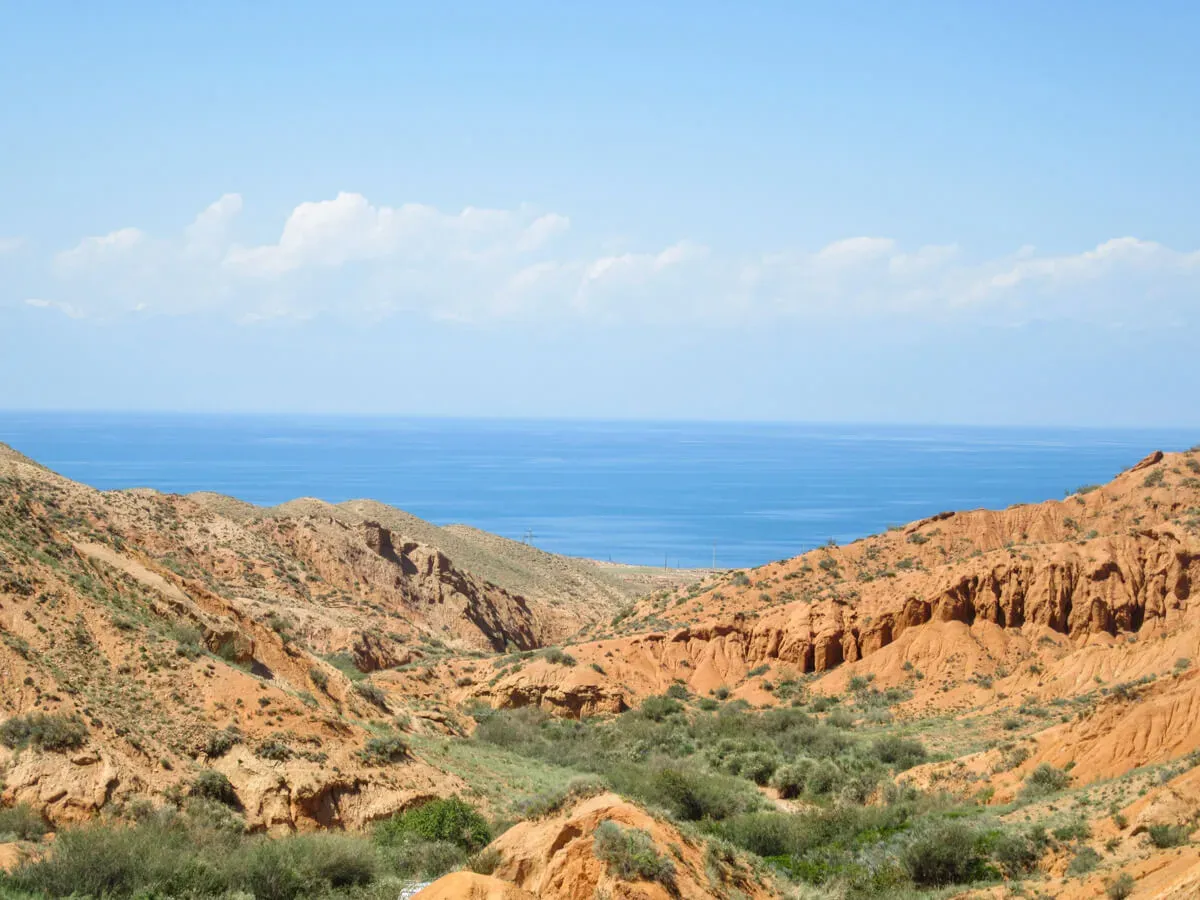 Panoramic view of Skazka Canyon red-orange rock formations with Lake Issyk-Kul in the background under a partly cloudy sky