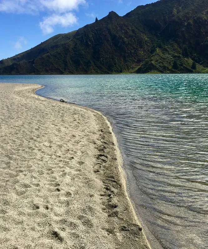 Sandy shoreline and brilliant turquoise waters of Lagoa do Fogo crater lake surrounded by lush green mountains