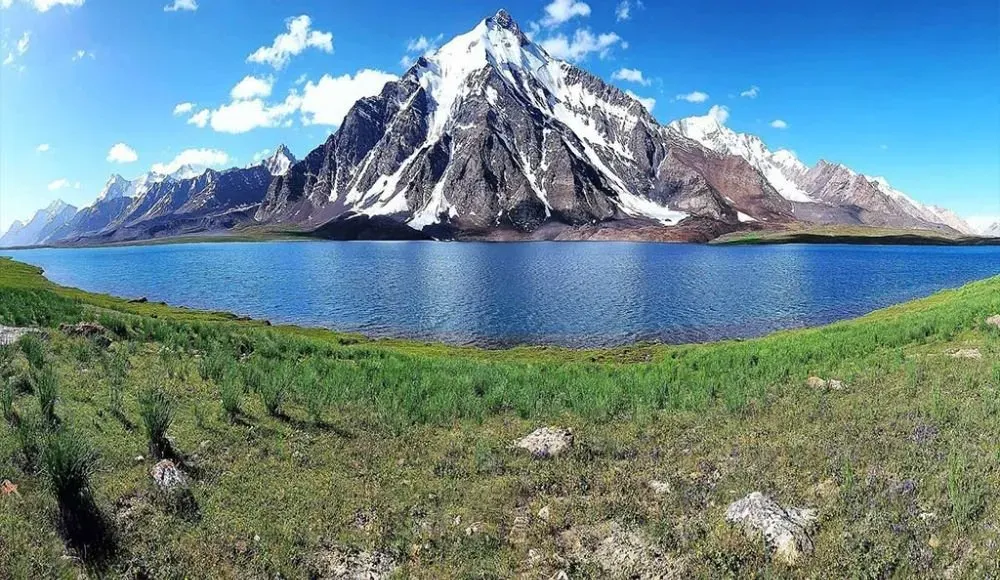 Snow-capped mountain rising above turquoise Karambar Lake in Pakistan with green grasslands