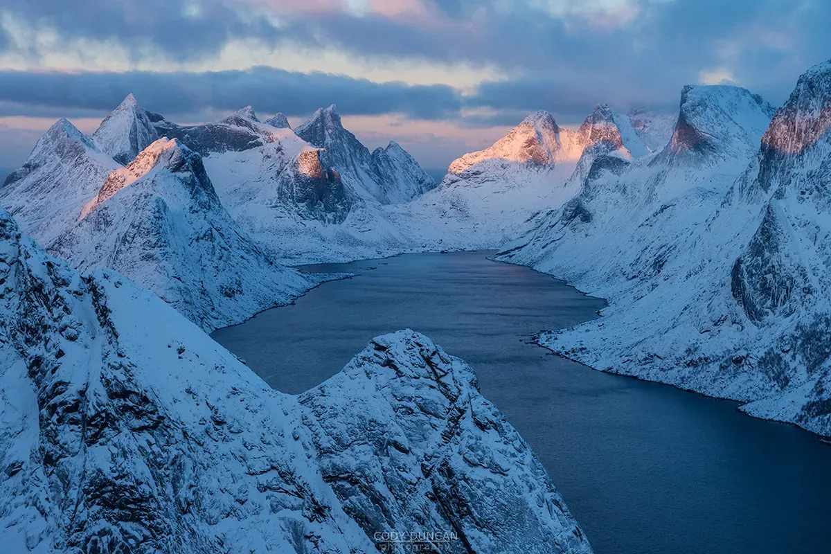Snow-covered Lofoten mountain peaks and calm fjord illuminated by soft pink and orange sunset light, Norway