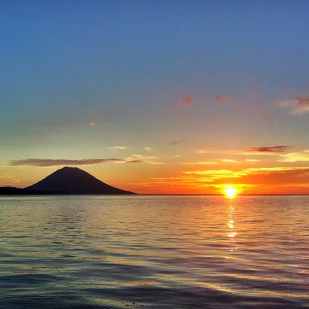 Sunset over Bunaken National Park Indonesia with silhouetted palm trees and Manado Tua volcano in the distance