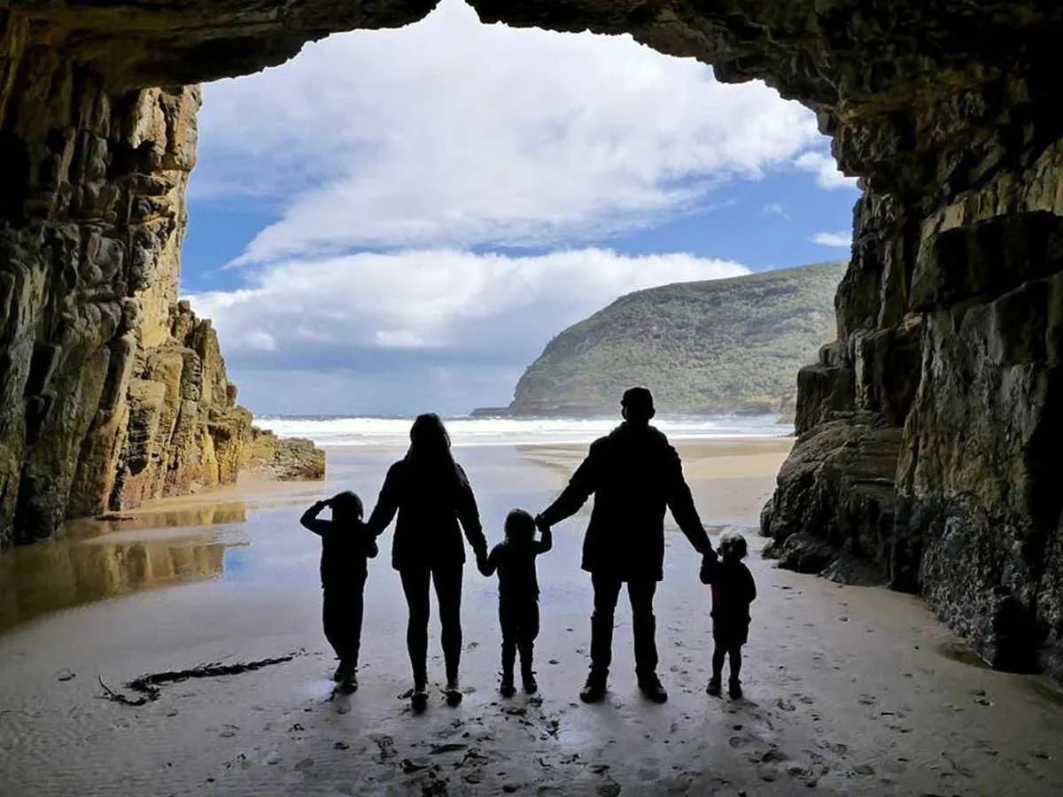 Visitors walk across a sandy beach inside Remarkable Cave with lush green headland visible through the opening