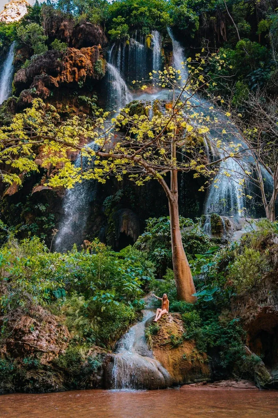Woman sitting on a rock at the base of Cascada El Aguacero waterfall surrounded by dense green vegetation