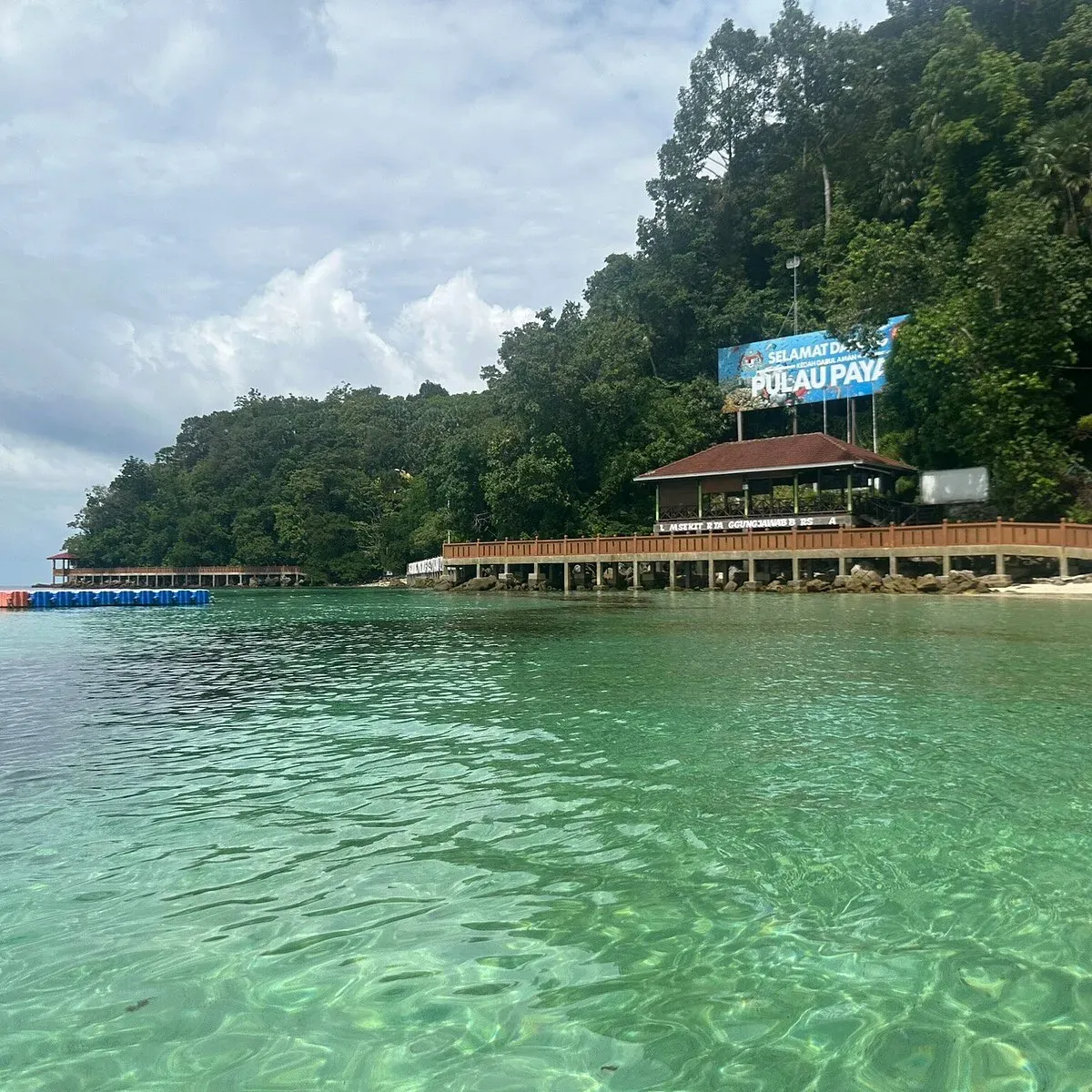 Wooden pier leading to Pulau Payar island with a welcome sign and small pavilion against a backdrop of lush green hills