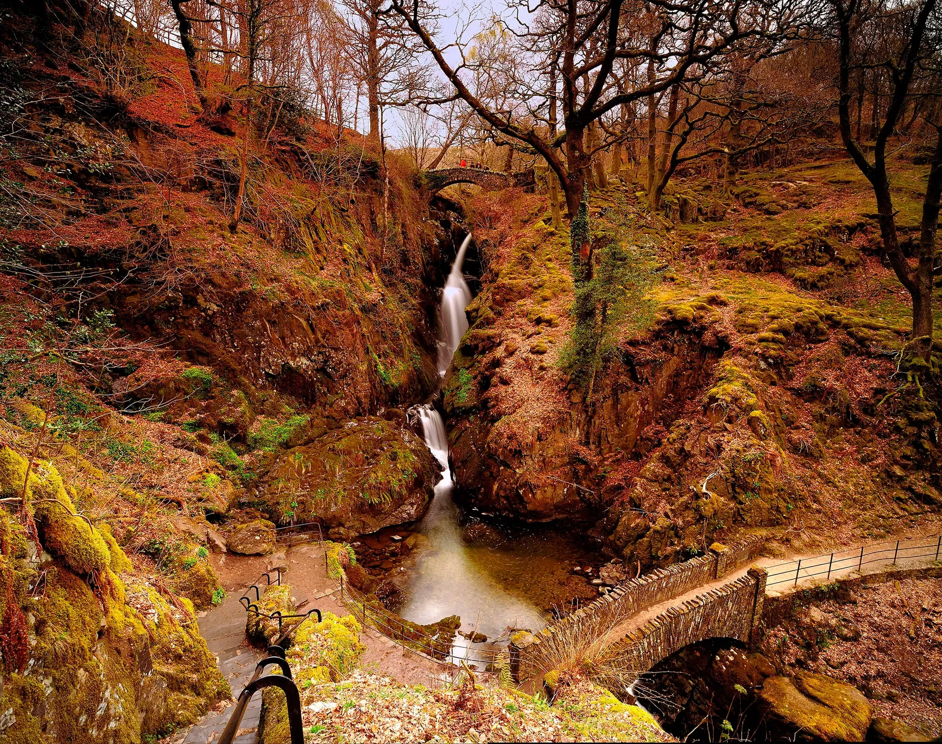 Aira Force waterfall in autumn with a stone footbridge crossing above and orange-leaved trees lining the gorge