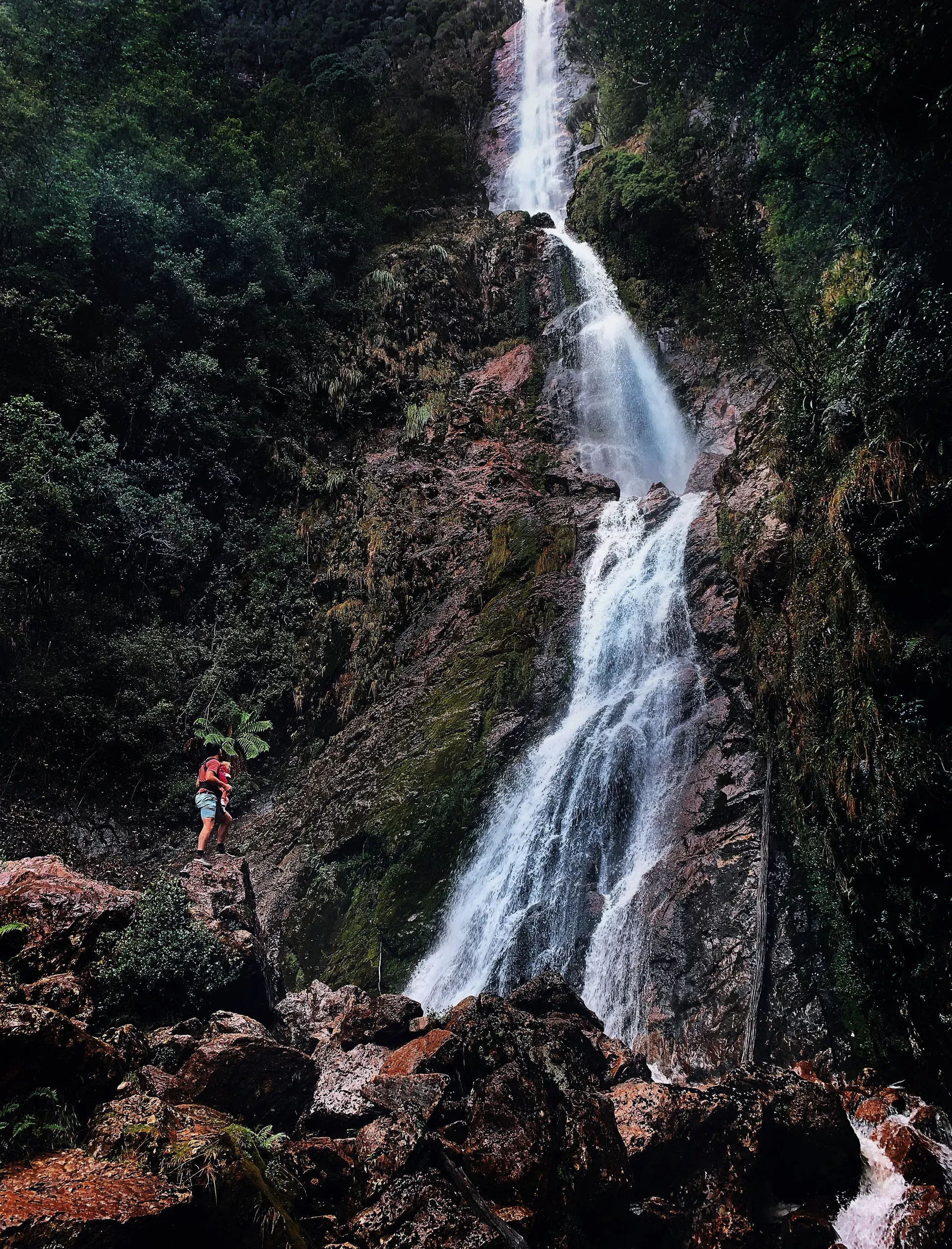Hiker at the base of Montezuma Falls Tasmania with the 104-metre waterfall behind them