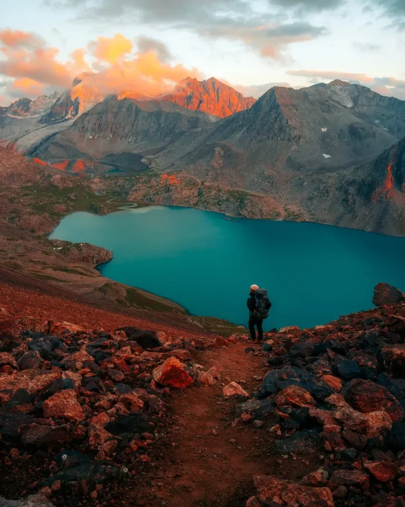 Hiker walking on rugged red rocky trail towards Ala-Kul Lake with towering mountains in golden light