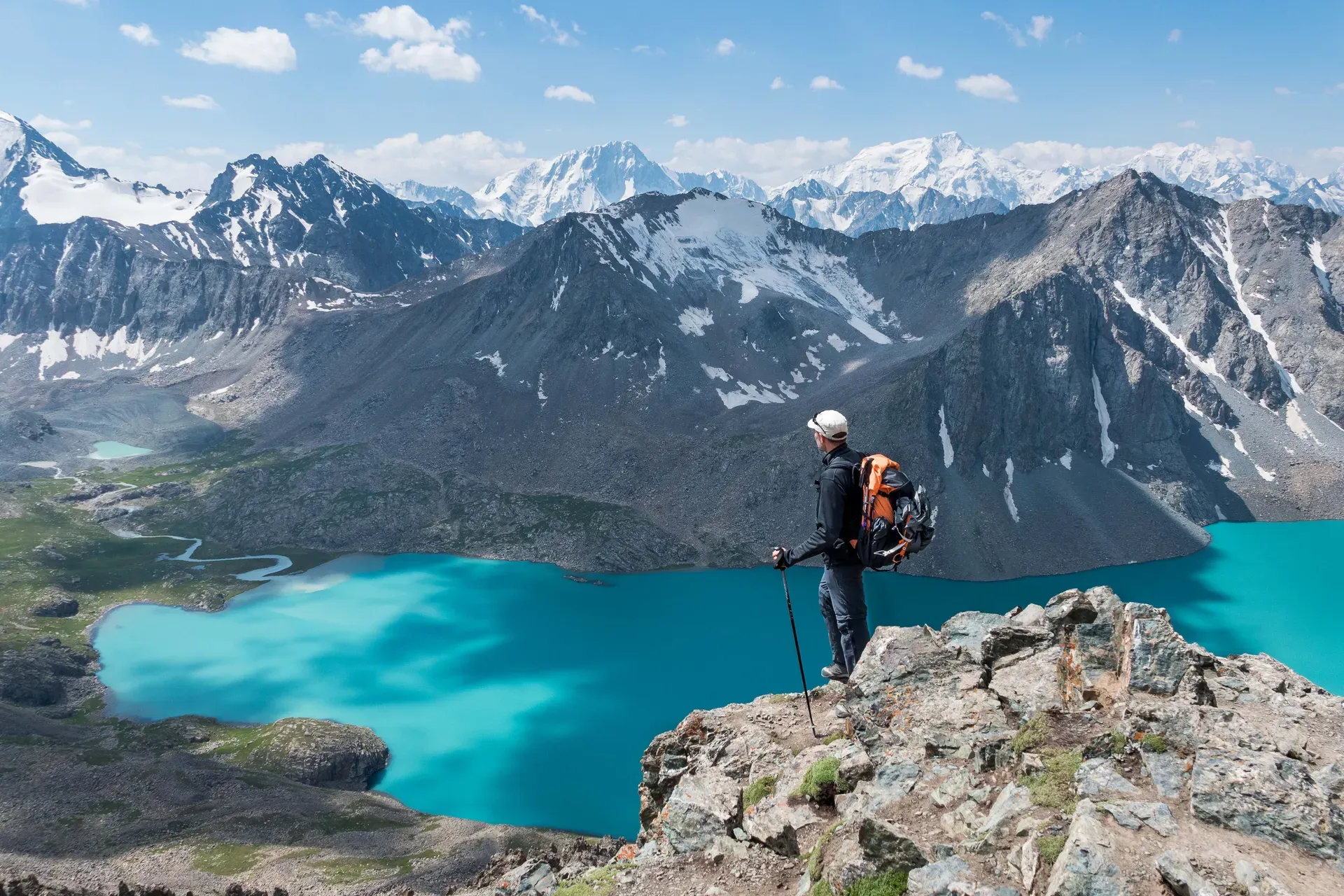 Hiker with backpack standing on rocky terrain overlooking vivid blue Ala-Kul Lake surrounded by snowy mountains