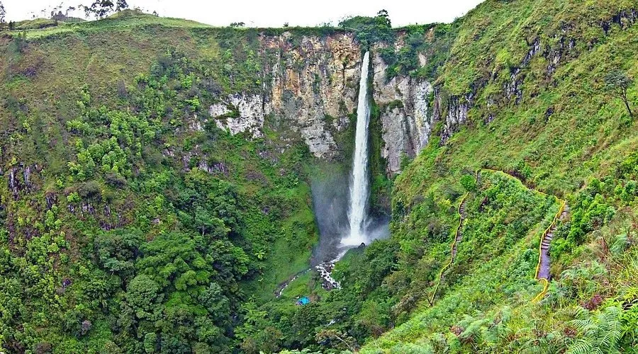 Sipiso Piso Waterfall plunging down a steep lush green hillside with a winding pathway toward Lake Toba