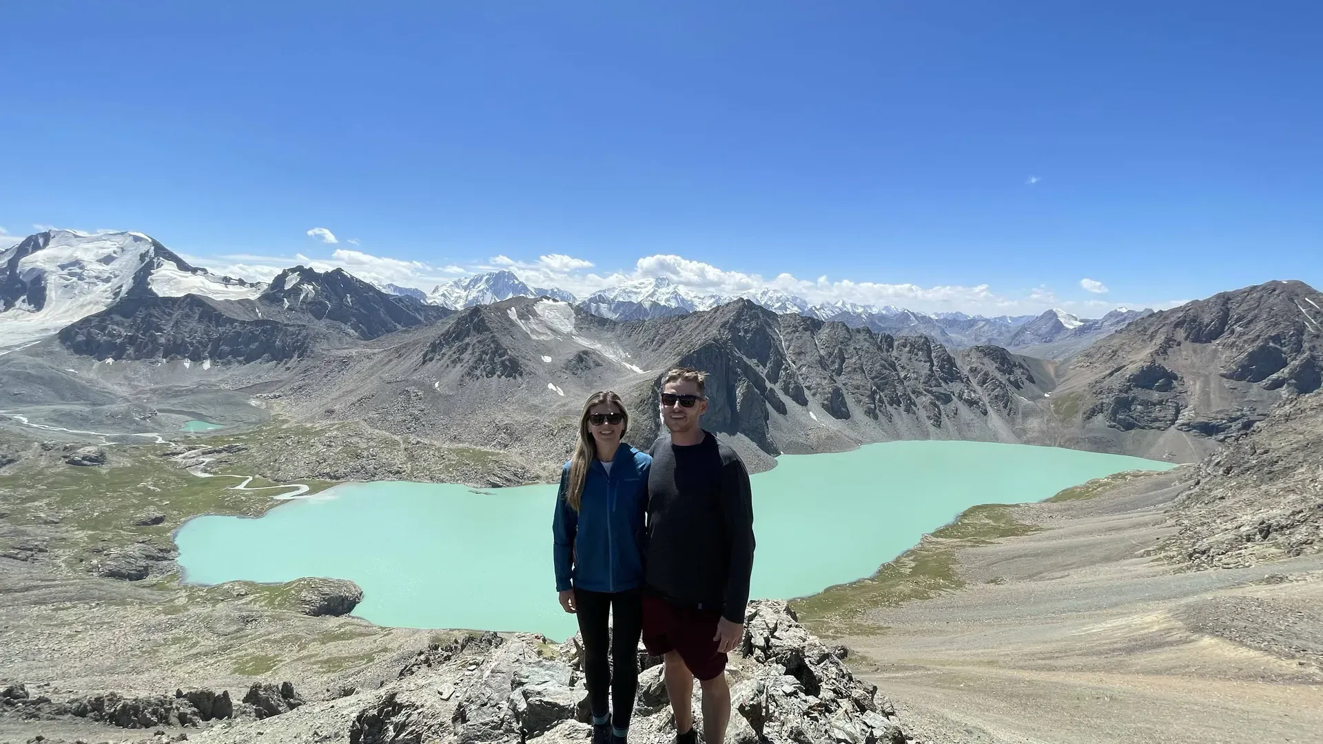 Smiling couple wearing sunglasses standing by turquoise Ala-Kul Lake with snow capped peaks behind them