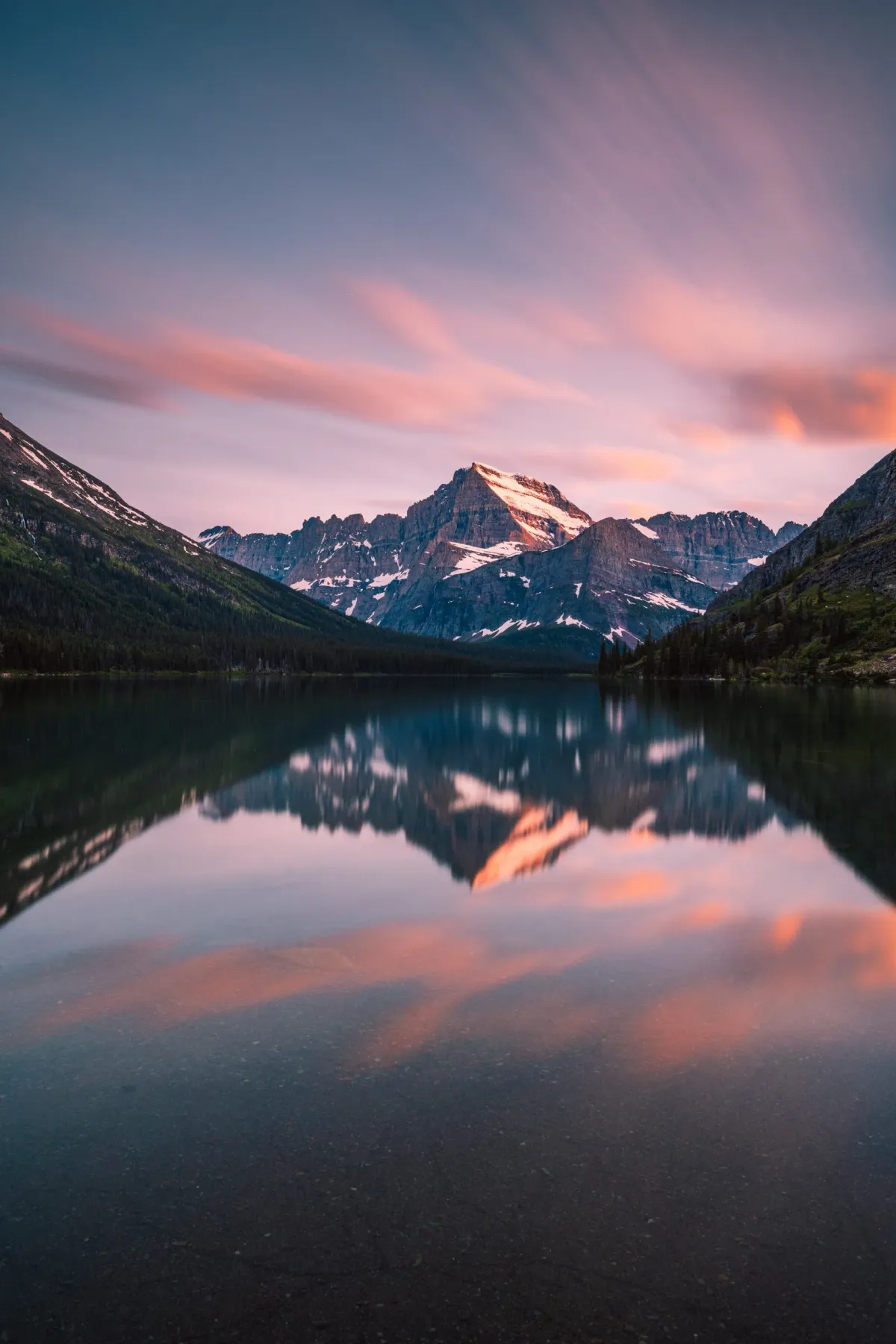 Snow-capped mountains reflected in calm Lake Josephine at sunset with pink clouds above in Glacier National Park