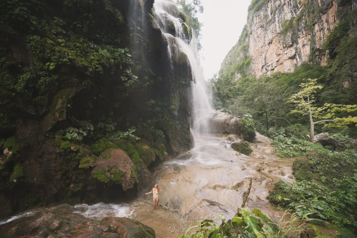 A person stands waist-deep in the cascading waters of Cascada El Aguacero surrounded by lush jungle in Chiapas, Mexico