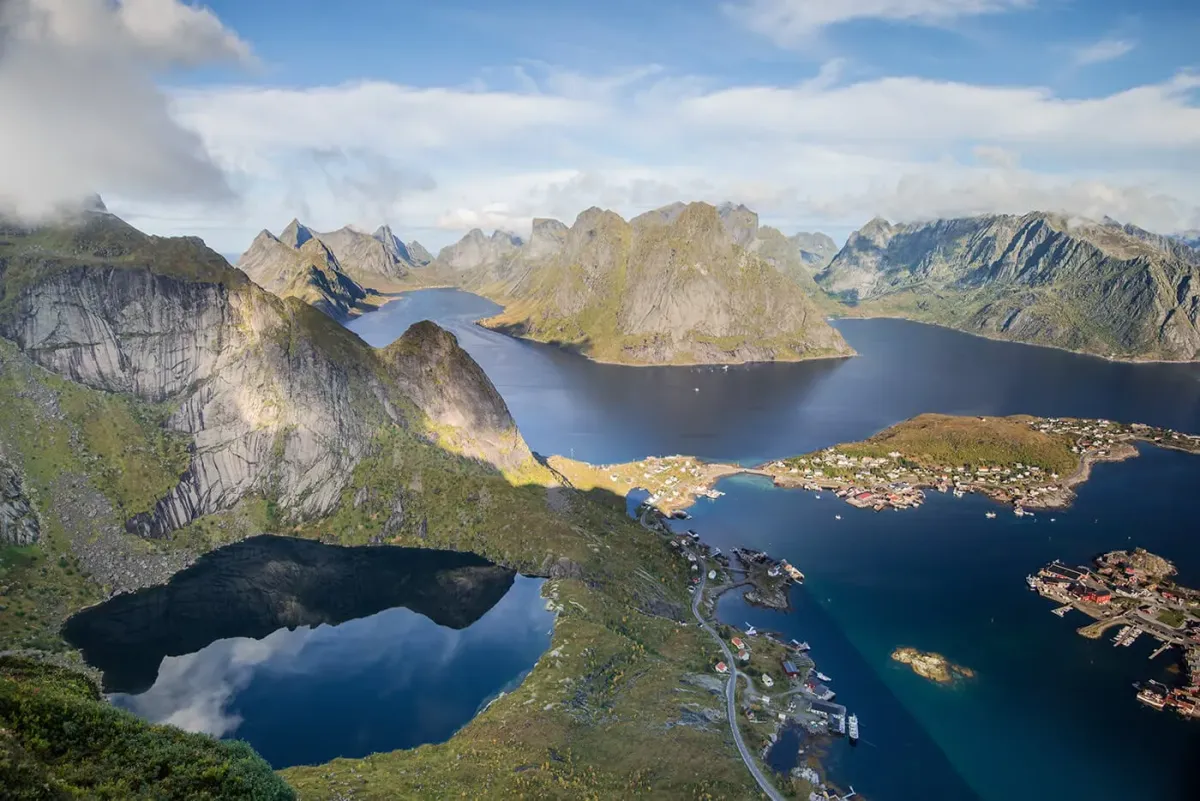 Panoramic view from Reinebringen summit overlooking Reinefjord, jagged Lofoten peaks, and the red rorbu fishing huts of Reine village, Norway