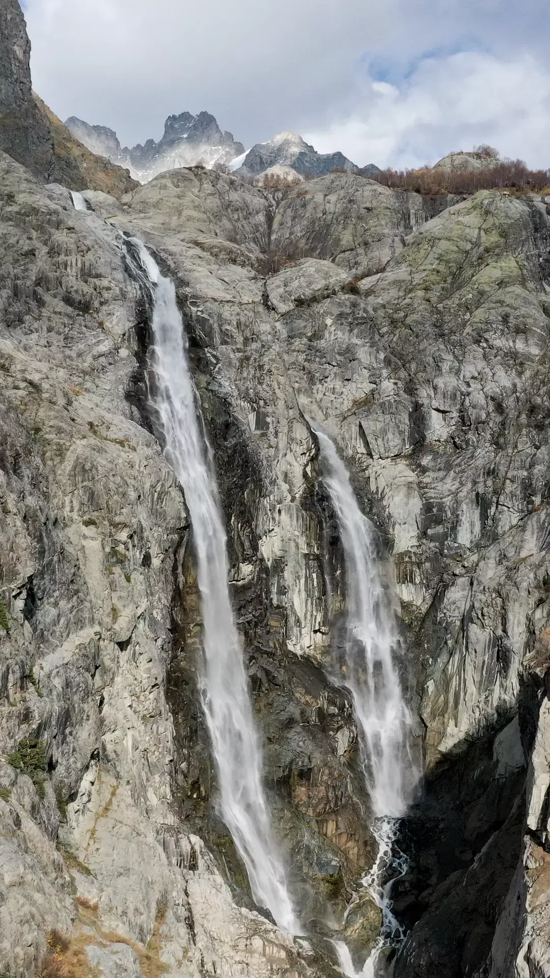 Shdugra Waterfall cascading down gray cliffs with snow-capped Caucasus mountains behind