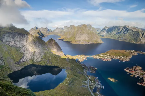 Panoramic view from Reinebringen summit overlooking Reinefjord, jagged Lofoten peaks, and the red rorbu fishing huts of Reine village, Norway