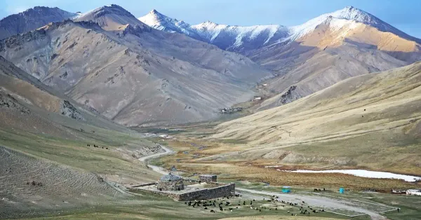 The Tash Rabat caravanserai, a 15th-century stone structure, set against green hills and the Tian Shan mountains in Kyrgyzstan under a bright sky